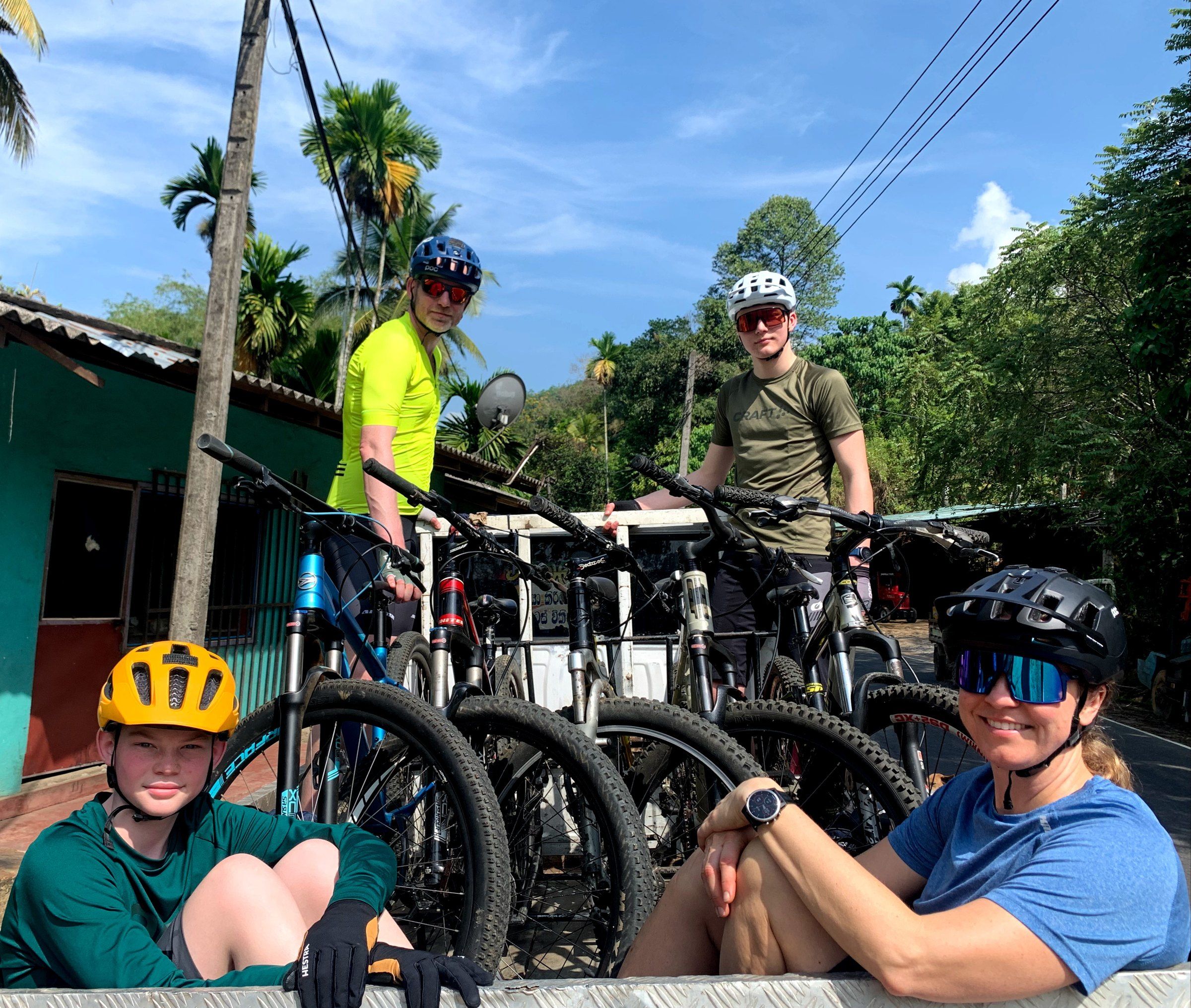 Four guests in helmets sit beside a truck loaded with mountain bikes