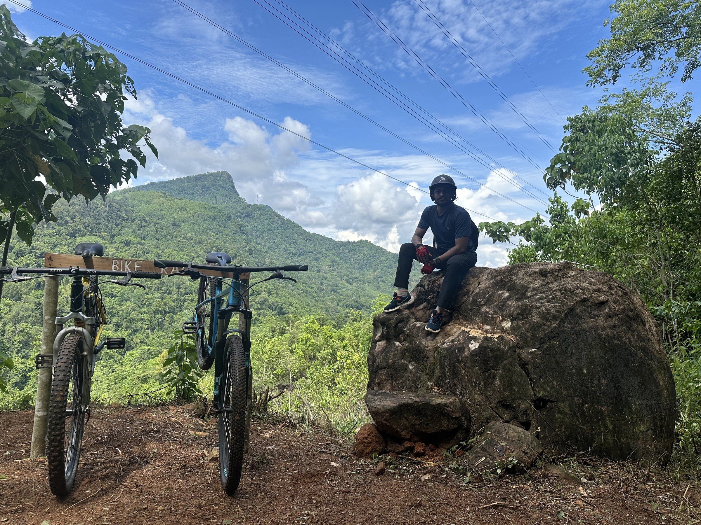 A rider sits on a large rock at the view point, two bikes parked beside him
