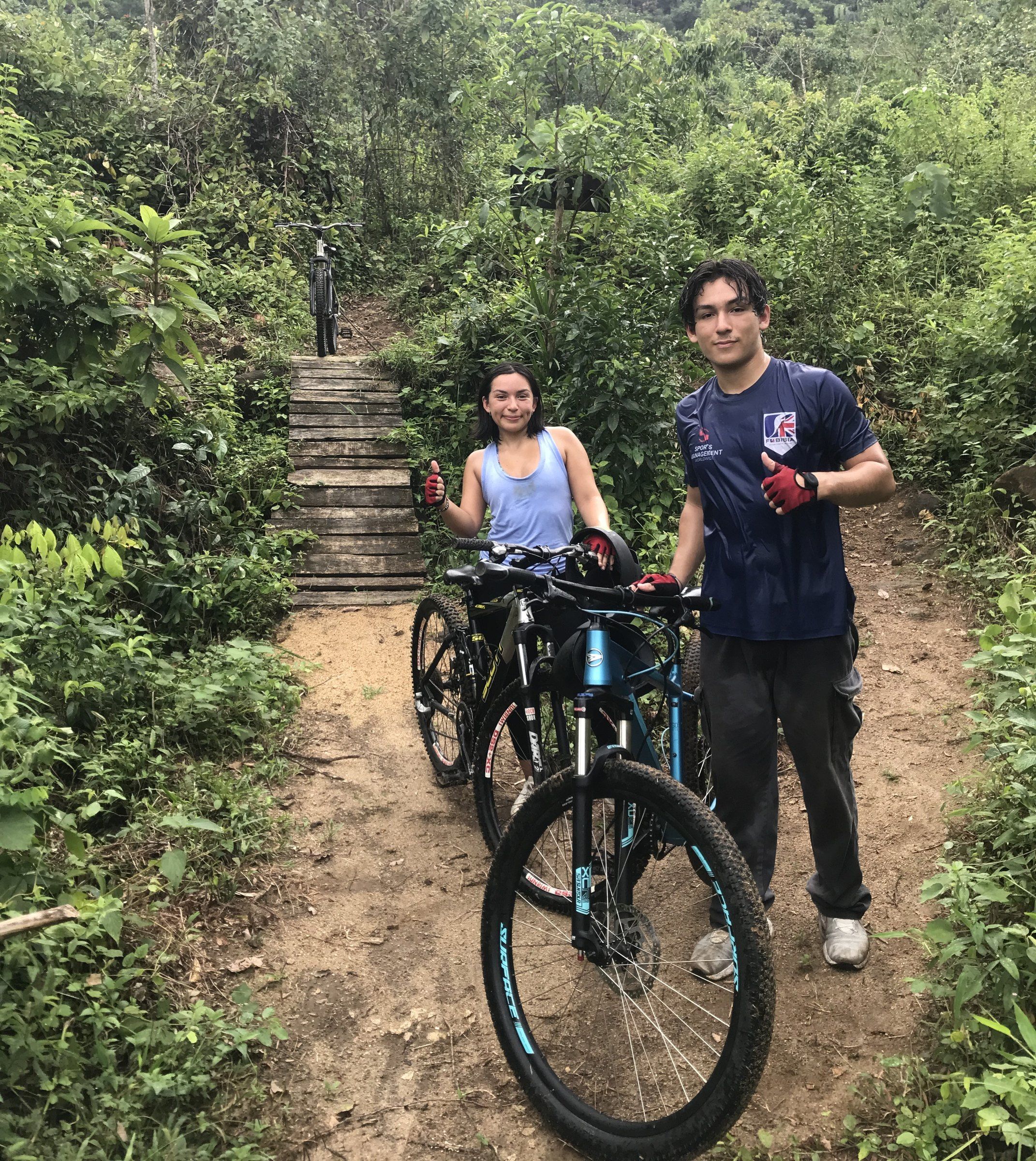 Two riders stand with their bikes on a dirt trail below stone steps