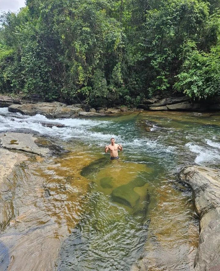 A guest stands waist-deep in a clear river pool between flat rocks, with jungle on the far bank